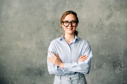 Confident young professional woman in a blue shirt and glasses smiling at the camera with arms crossed. Perfect for business profiles, leadership, and professional success themes