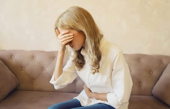 Young woman put hands on abdomen suffering from stomach ache griping bellyache feel bad with pain sitting on a couch at home