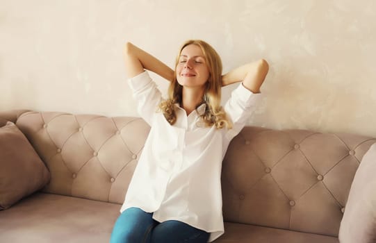 Happy relaxed young woman enjoying at home, satisfied girl resting with closed eyes and folded hands behind the head sitting on the couch in living room