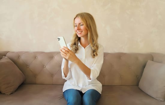 Happy smiling young woman looking at phone, girl with smartphone sitting on the couch in interior room at home