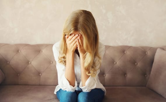 Upset sad crying woman covering her eyes and face with her hands while experiencing mental or physical suffering, fatigue sitting on the couch in room at home