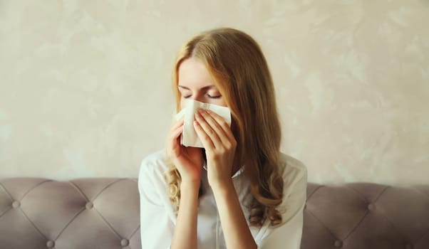 Sick exhausted young woman sneezing blow nose using tissue, unhealthy suffering girl feels unwell sitting on the couch at home