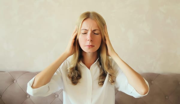 Exhausted tired woman touching head, suffering from headache, stress, fatigue, temples pain, migraine, loud noise, tension sitting on the couch at home