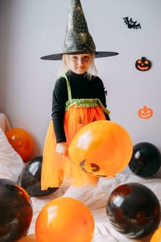 Children's Halloween - a girl in a witch hat and a carnival costume with airy orange and black balloons at home. Ready to celebrate Halloween