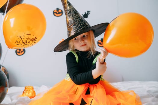 Children's Halloween - a girl in a witch hat and a carnival costume with airy orange and black balloons at home. Ready to celebrate Halloween.