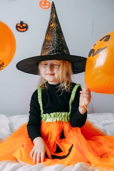 Children's Halloween - a girl in a witch hat and a carnival costume with airy orange and black balloons at home. Ready to celebrate Halloween.