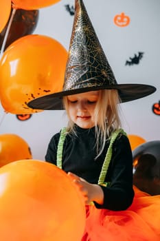 Children's Halloween - a girl in a witch hat and a carnival costume with airy orange and black balloons at home. Ready to celebrate Halloween.
