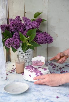 female hand cutting cake decorated with lilac flowers near vase with lilac branches on wooden table emphasizing floral and rustic lifestyle, delicate still life,