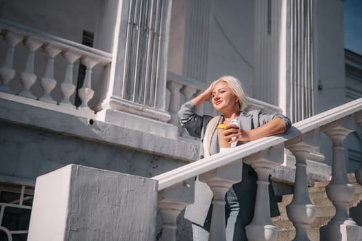 A woman is standing on a white railing with a cup of coffee in her hand. She is wearing a gray jacket and blue jeans. Concept of relaxation and leisure