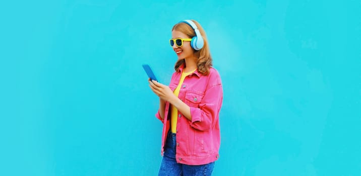 Portrait of stylish modern happy smiling young woman in headphones listening to music with smartphone wearing pink jacket on vivid blue background