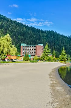 Sand shore of the lake leading to resort building on green mountain and blue sky background