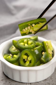 A food photograph of a bowl of sliced peppers and a pair of chopsticks holing a slice.