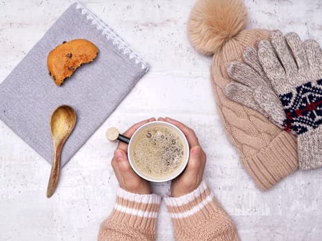 A flatlay photo of a woman's hands holding a cup of coffee, a wooden spoon, cookie, gloves, and a hat on the side.