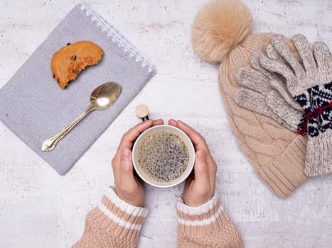 A flatlay photo of a woman's hands holding a cup of coffee, a wooden spoon, cookie, gloves, and a hat on the side.
