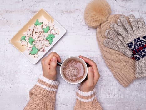 A flatlay photo of a woman's hands holding a cup of hot cocoa, animal crackers, gloves, and a hat on the side.