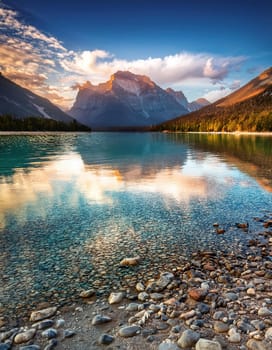 A stunning image of a crystal-clear lake with smooth, colorful pebbles lining the shore. The water reflects the vibrant blue sky and fluffy clouds above, creating a truly breathtaking scene.This mesmerizing photo showcases the beauty of nature with a close-up of smooth, colorful pebbles glistening in the crystal-clear water of a serene lake. The surrounding landscape completes the picture with lush green mountains and a bright blue sky.