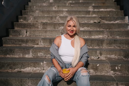 A blonde woman is sitting on a set of stairs, holding a cup of coffee. She is wearing a white tank top and jeans
