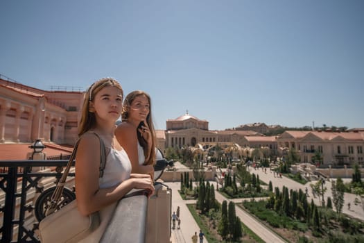 Two young women stand on a balcony overlooking a sprawling city park with buildings in the background. They are looking out at the scene.
