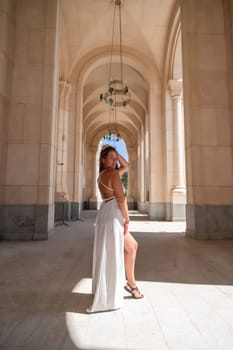 A woman in a white dress stands in a stone hallway with arches, sunlight illuminating the scene.