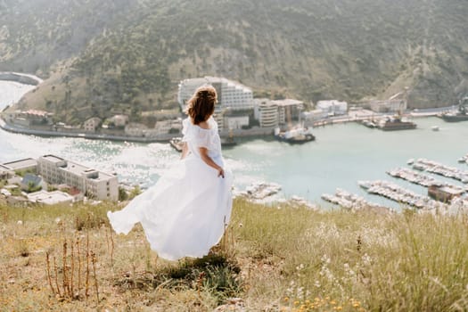 Happy woman in a white dress and hat stands on a rocky cliff above the sea, with the beautiful silhouette of hills in thick fog in the background