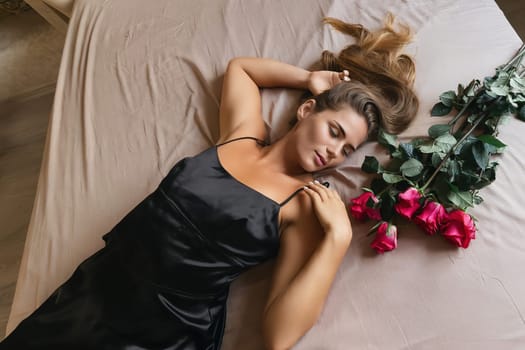 A woman is laying on a bed with a bouquet of red roses next to her. Concept of romance and intimacy, as the woman is surrounded by the flowers and he is in a relaxed and comfortable position