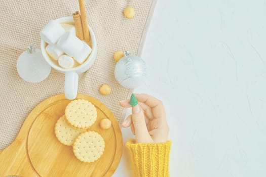 Woman in a yellow sweater drinks hot cocoa with marshmallow and cinnamon rolls in a white mug and cookies and a figure of a Christmas tree