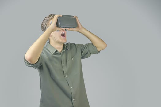 Young man in shirt enjoys and amazed by virtual reality glasses in front of light grey background