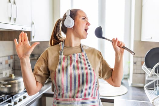 Woman wearing headphones and apron sings and dances in kitchen. Casual indoor lifestyle photography. Joy of cooking and music concept for design and print.