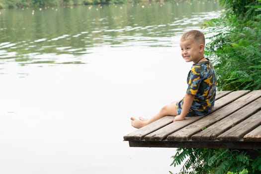 Fishing concept. A small handsome boy, with emotional facial expressions, 4 years old, sits on a wooden bridge by the river, against the backdrop of green plants and breathes fresh air. Relax. Place for text. Copy space. Vacations, Village.