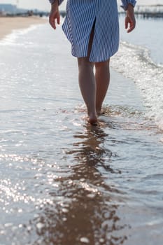 young woman barefoot walks along the sandy beach by the sea at sunset, plunging her feet into the waves and sand, pleasant relaxation on summer vacation