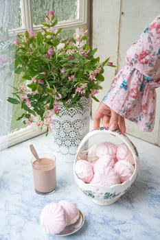 female hand holding full basket of vanilla marshmallows near vase with pink flowers, home cooking and country life concept,