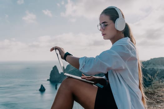 Digital nomad, woman in the hat, a business woman with a laptop sits on the rocks by the sea during sunset, makes a business transaction online from a distance. Freelance, remote work on vacation.