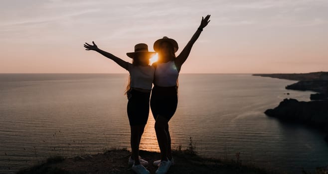 Two women wearing hats stand side-by-side against the silhouette of a cliff with the sea and a sunset behind them.