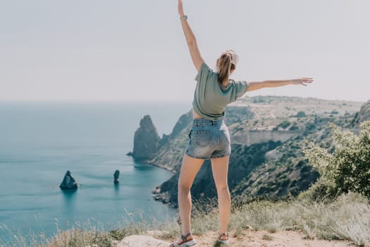 A young woman, wearing denim shorts and a green shirt, stands on a clifftop with her arms raised, looking out at a stunning coastal view. The photo captures a moment of pure joy and freedom, set against a backdrop of blue sky and water.