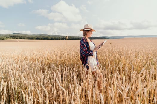 A young woman in a hat and plaid shirt stands in a wheat field and uses a tablet.