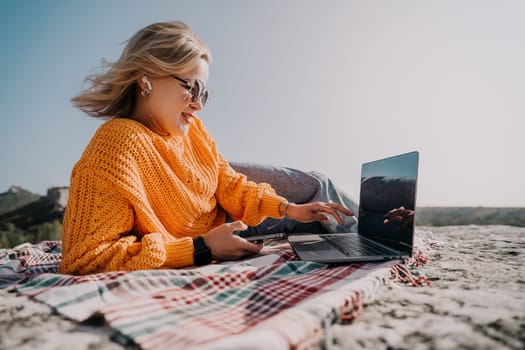 A woman with blonde hair, wearing sunglasses and a yellow sweater, works on a laptop while enjoying the outdoors.