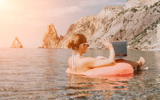 Woman freelancer works on laptop swimming in sea on pink inflatable ring. Pretty lady typing on computer while floating in the sea on inflatable donut at sunset. Freelance, remote work on vacation