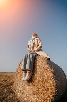 A young woman wearing a beige trench coat and blue jeans sits on a large hay bale, looking up at the clear blue sky.