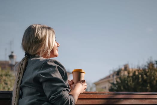A woman sits on a bench with a cup of coffee in her hand. She is smiling and enjoying the view