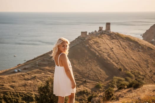 A blonde woman stands on a hill overlooking the ocean. She is wearing a white dress and she is enjoying the view