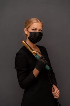 A woman in a black dress holding a hair curler. She is wearing a mask. Concept of caution and responsibility in the face of a pandemic