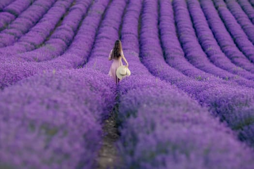 A woman is walking through a field of purple flowers. She is wearing a white hat and a pink dress. The scene is peaceful and serene
