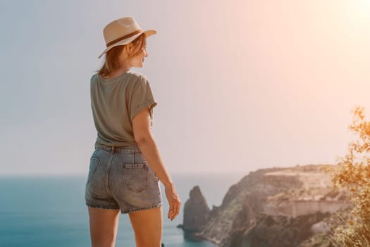 A young woman wearing a straw hat and a green shirt stands on a cliff, gazing out at the ocean.