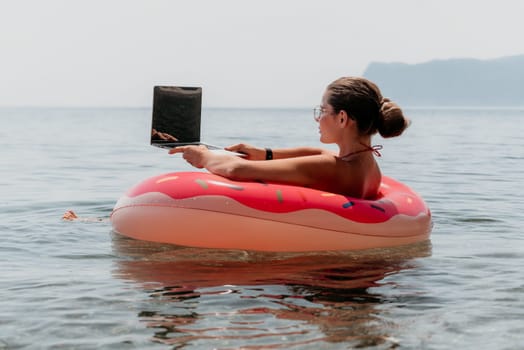 Woman freelancer works on laptop swimming in sea on pink inflatable ring. Pretty lady typing on computer while floating in the sea on inflatable donut at sunset. Freelance, remote work on vacation