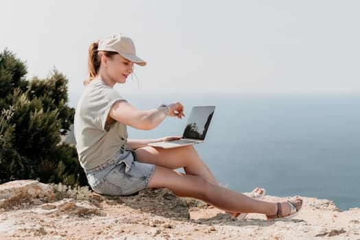 A young woman in a cap and casual clothes sits on a cliffside, using a laptop computer with the sea in the background.