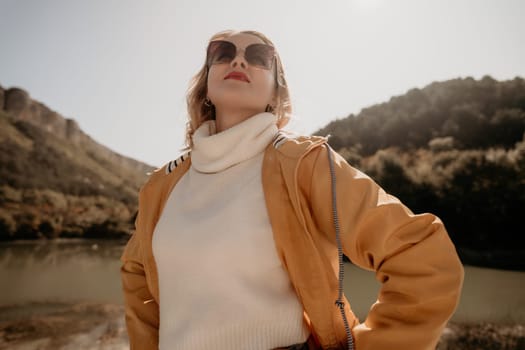 A young woman in a yellow jacket and sunglasses stands by a lake, with hills in the background and a blue sky overhead.