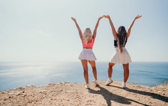 Two women stand on a cliffside with their backs to the camera, their arms raised in a triumphant pose. The ocean stretches out before them under a clear blue sky.