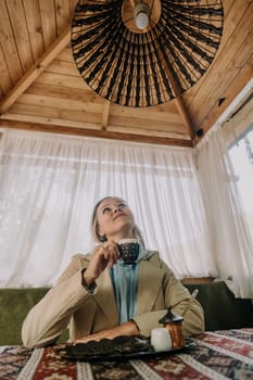 A woman dressed in a beige jacket enjoys Turkish coffee in a cozy cafe, gazing up at the intricate ceiling lamp.