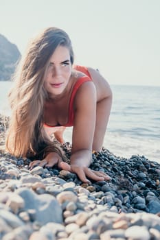 A young woman wearing a red swimsuit crawls on a pebble beach while looking at the camera.