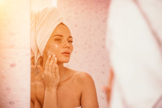 Young beautiful woman using face cream moisturizing lotion after bath. Pretty attractive girl in a towel on her head stands in front of a mirror in a home bathroom. Daily hygiene and skin care.